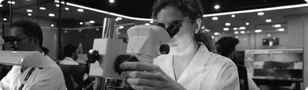 A high-contrast black and white photograph of an SGL's gemologist using a professional laboratory microscope for precise diamond grading and stone inspection.